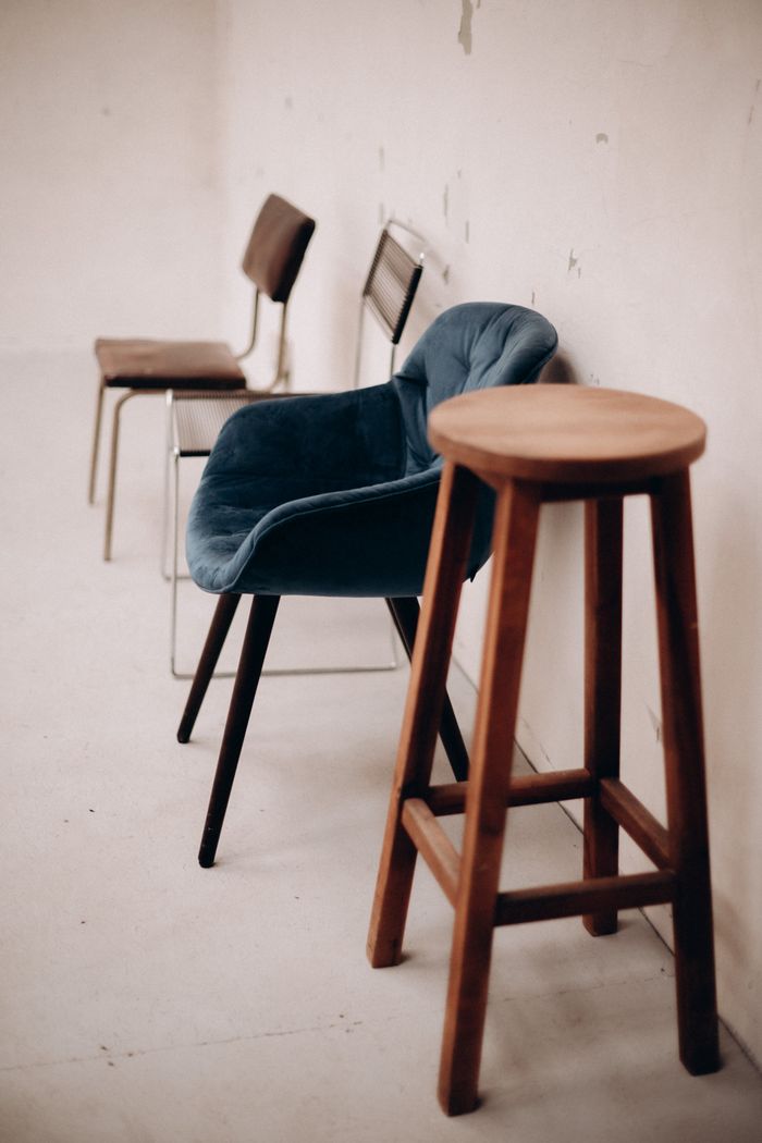 A minimalist room featuring a wooden stool and various chairs against a white wall.